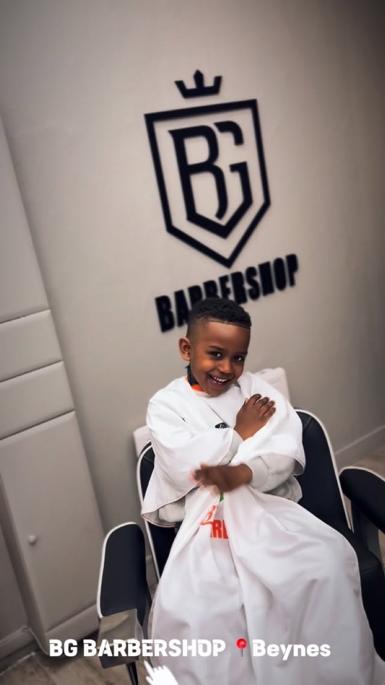 Jeune enfant souriant, installé dans le fauteuil avec la cape blanche, sous le logo BG Barbershop à Beynes